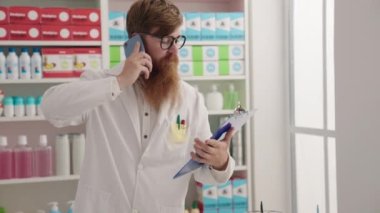 Young redhead man pharmacist talking on smartphone reading document at pharmacy