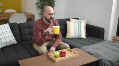 Genç kel adam kahvaltı yapıyor. Evdeki koltukta oturuyor.