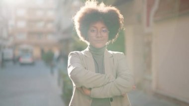 Afro-Amerikalı kadın gülümsüyor. Kollarını kavuşturup caddede duruyor.