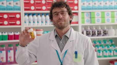 Young hispanic man pharmacist smiling confident holding pills bottle at pharmacy