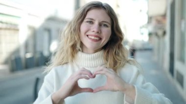 Young blonde woman doing heart gesture with hands over eyes at street