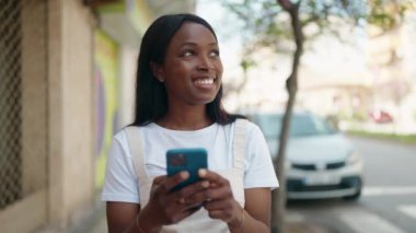 Genç Afro-Amerikalı kadın sokakta akıllı telefon kullanarak gülümsüyor.