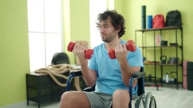 Young hispanic man sitting on wheelchair training with dumbbells at rehab clinic