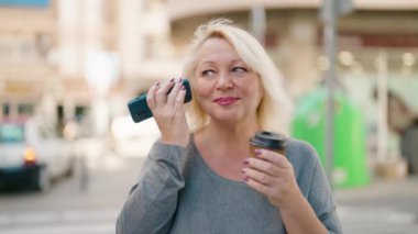 Middle age blonde woman listening audio message by the smartphone drinking coffee at street