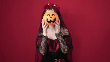 Young blonde woman wearing katrina costume holding halloween pumpkin basket over face over isolated red background