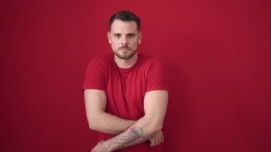 Young caucasian man standing with serious expression taking t shirt off over isolated red background