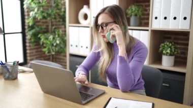 Genç sarışın iş kadını bilgisayarını kullanarak ofiste akıllı telefondan konuşuyor.
