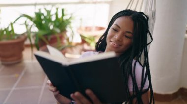 Afro-Amerikan kadın kitap okuyor. Hamakta yatıyor.