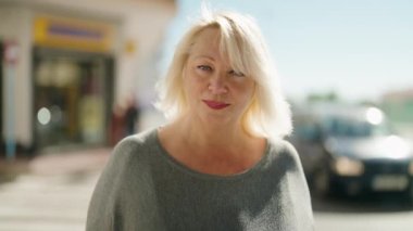 Orta yaşlı sarışın kadın sokakta kendinden emin bir şekilde gülümsüyor.