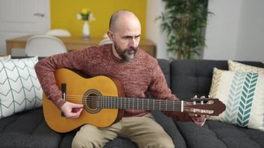 Genç kel adam evdeki kanepede oturup klasik gitar çalıyor.