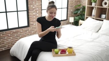 Young beautiful hispanic woman making photo to healthy breakfast by smartphone at bedroom