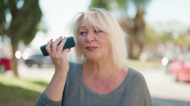 Orta yaşlı sarışın kadın gülümsüyor. Parktaki akıllı telefondan kendine güvenen sesli mesaj dinliyor.