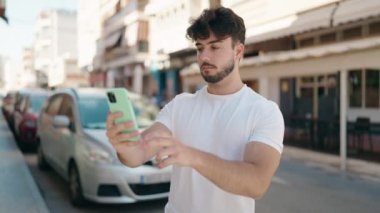 Young hispanic man smiling confident making selfie by the smartphone at street