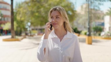 Genç sarışın kadın gülümsüyor. Parktaki akıllı telefondan konuşuyor.