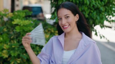Young beautiful hispanic woman smiling confident using dollars as a handfan at street