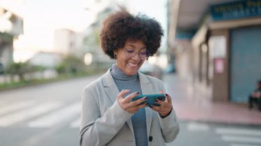 Afro-Amerikalı kadın gülümsüyor. Sokakta akıllı telefondan videolar izliyor.