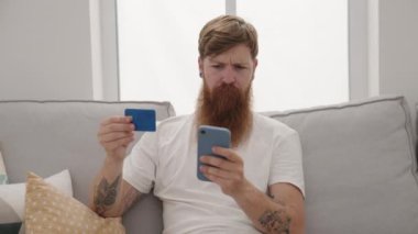 Young redhead man using smartphone and credit card sitting on sofa at home