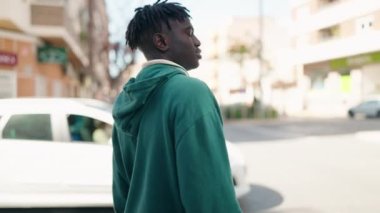 Afro-Amerikalı adam gülümsüyor. Kendine güveni tam. Sokakta çapraz hareketlerle duruyor.