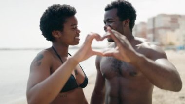 Afrika kökenli Amerikalı erkek ve kadın mayo giyen çift deniz kenarında kalp sembolü yapıyorlar.