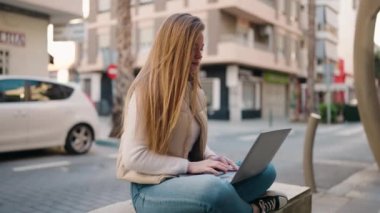 Young blonde woman using laptop sitting on bench at street