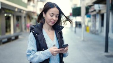 Genç, güzel, İspanyol bir kadın sokakta akıllı telefon kullanarak gülümsüyor.