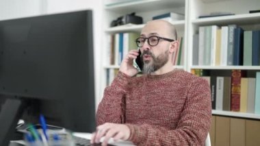 Genç, kel bir öğrenci üniversite sınıfında bilgisayar kullanarak akıllı telefondan konuşuyor.