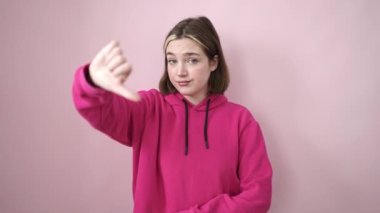 Genç sarışın kadın negatif jest yapıyor, pembe arka plan üzerinde başparmakları var.