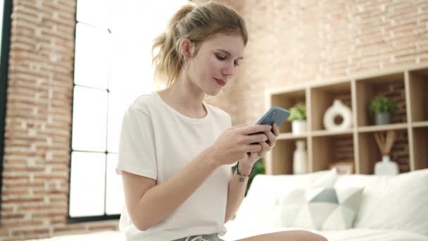 年轻的金发女人坐在卧室的床上用智能手机 — 图库视频影像