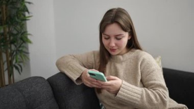 Akıllı telefon kullanan genç sarışın kadın evde iyi bir jest yapıyor.