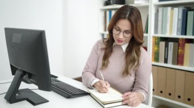 Young beautiful hispanic woman student using computer writing on notebook at library university