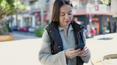 Genç, güzel, İspanyol bir kadın sokakta akıllı telefon kullanarak gülümsüyor.