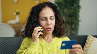 Orta yaşlı İspanyol kadın elinde kredi kartıyla akıllı telefondan konuşuyor.
