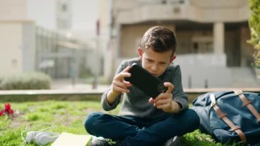 Parktaki çimlerin üzerinde oturan sarışın çocuk akıllı telefondan video oyunu oynuyor.