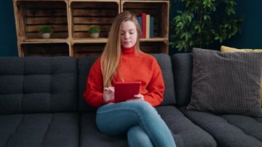 Young blonde woman using touchpad sitting on sofa at home