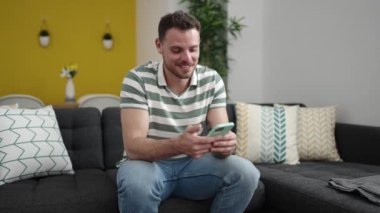 Young caucasian man smiling using smartphone sitting on the sofa at home