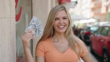 Young blonde woman smiling confident holding dollars at street