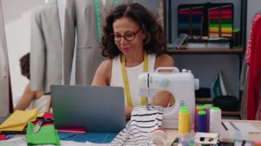 Middle age woman tailor smiling confident using laptop at clothing factory