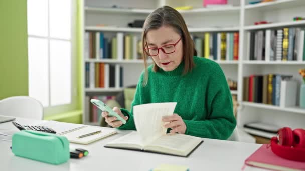 在图书馆用智能手机阅读一本书的成熟的灰发女学生 — 图库视频影像