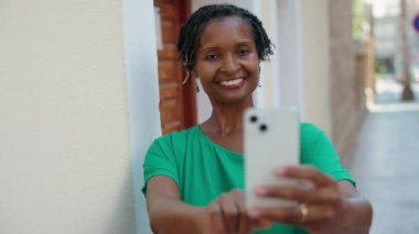 Orta yaşlı Afro-Amerikalı kadın gülümsüyor. Kendine güveni tam. Sokakta akıllı telefondan selfie çekiyor.