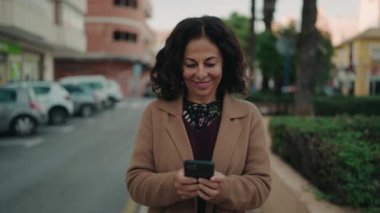 Orta yaşlı İspanyol kadın sokakta akıllı telefon kullanarak gülümsüyor.