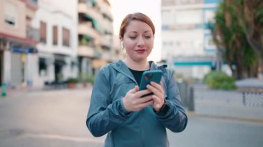 Spor kıyafetleri giyen genç kızıl saçlı kadın sokakta güçlü jestler yaparak selfie çekiyor.