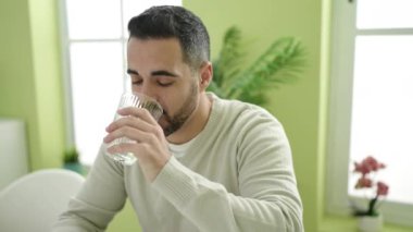 Young hispanic man drinking water sitting on table at home