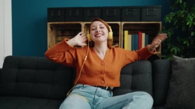 Young redhead woman listening to music and dancing sitting on sofa at home