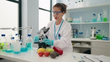 Laboratuvarda mikroskop kullanan orta yaşlı kadın bilim adamı üniforması giyiyor.