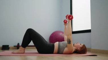 Genç kızıl saçlı kadın spor merkezinde halterleri kullanarak kendine güvenen bir şekilde gülümsüyor.