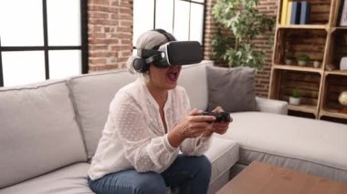 Orta yaşlı, kır saçlı, video oyunu oynayan bir kadın. Evde video gözlüğü ve joystick kullanıyor.