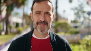 Orta yaşlı bir adam gülümsüyor. Parkta kollarını kavuşturmuş duruyor.