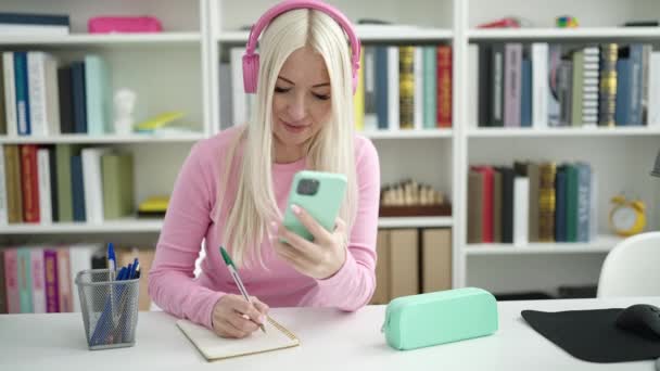 Νεαρή Ξανθιά Φοιτήτρια Χρησιμοποιώντας Smartphone Και Ακουστικά Γράφοντας Στο Σημειωματάριο — Αρχείο Βίντεο