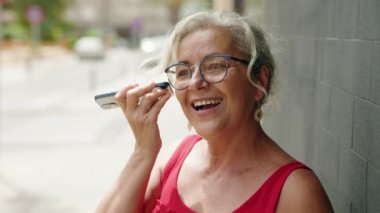 Orta yaşlı, kır saçlı, gülümseyen, kendine güvenen, akıllı telefondan sesli mesaj dinleyen bir kadın.