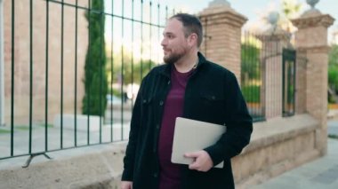 Genç ve cüsseli adam gülümsüyor. Elinde dizüstü bilgisayarla sokakta dans ediyor.